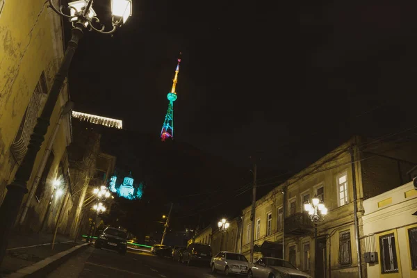 Mtatsminda street in Tbilisi at night with standing cars and traditional neighborhood houses  and Tv tower on the background lit with green red yellow light on 11th of March. 2020