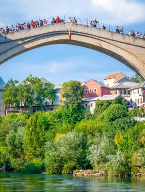 Mayo giymiş bir erkek, Mostar 'daki Most Musala Köprüsü' nden nehre atlıyor. Turistler bakıyor ve fotoğraf çekiyor. Bosna 'da turizm ve faaliyetler.