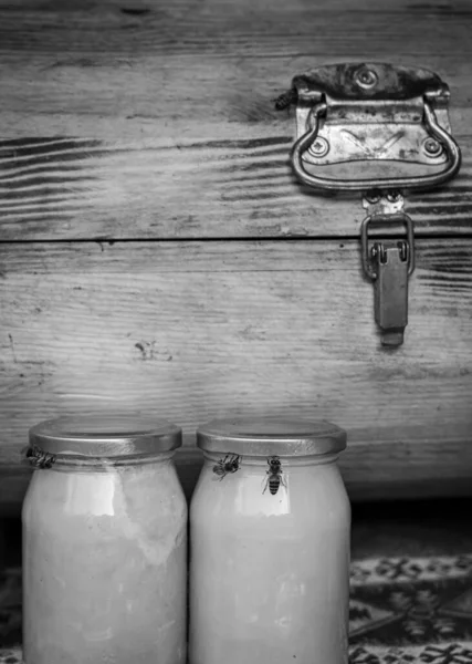 B&w image of two honey jars and bees trying to get inside them with old vintage storage box in the background. Storage and industry of bee keeping.