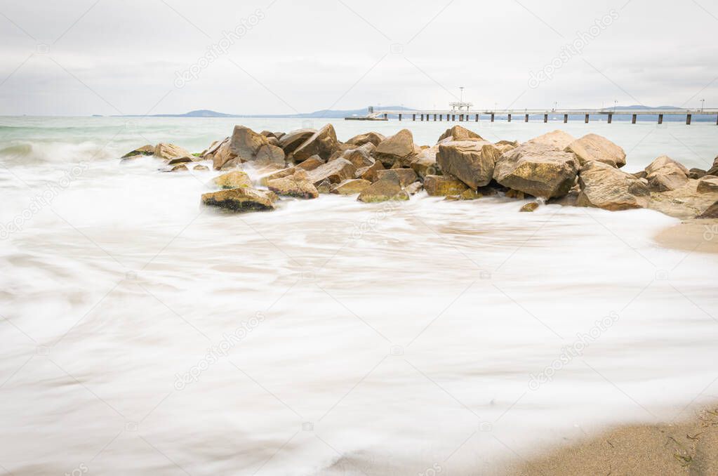 Rocks on the beach with silky sea water and Burgas bridge in the background. Fine art image Bulgaria. Travel destination 2020.
