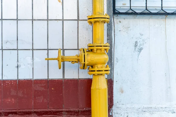 Big yellow gas valve on the pipe near balcony and wall with tiles. Valve with flywheel on the pipe allowed to distribute gas and turn it off if needful. On vertical pipe located flanges and reducer.