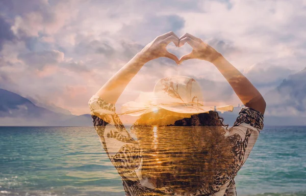 Double exposure of woman doing heart shape gesture and sunset on lakescape — Stock Photo, Image