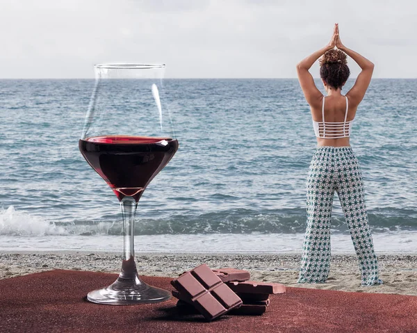 Copo de vinho tinto com chocolate e mulher praticando ioga perto do mar — Fotografia de Stock