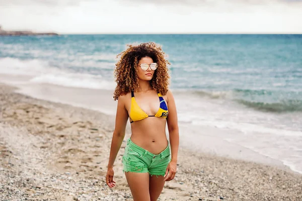 Retrato de mujer en bikini, pantalones cortos y gafas de sol y caminando por la playa — Foto de Stock