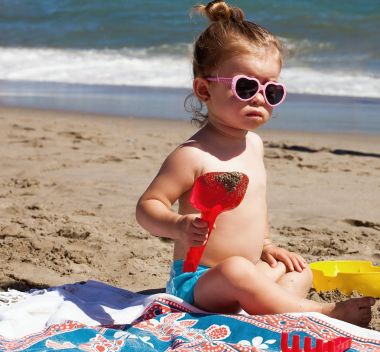Sahilde oturup, kalp şeklinde güneş gözlüğü takıyor, kumla oynayan küçük kız