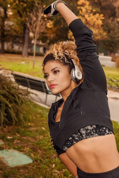 Retrato jogger haciendo estiramiento en el parque otoñal mientras usa auriculares y ropa deportiva —  Fotos de Stock