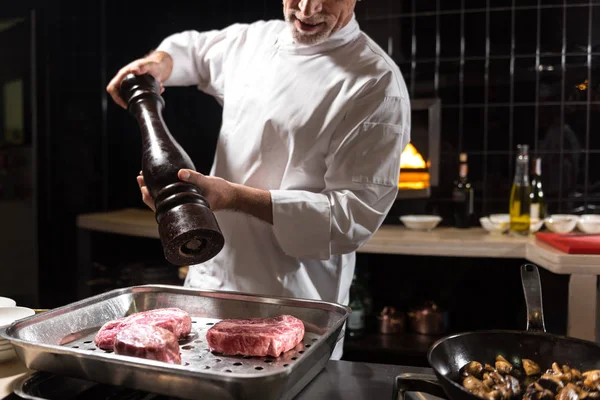 Bearded smiling chef putting salt on steaks — Stock Photo, Image