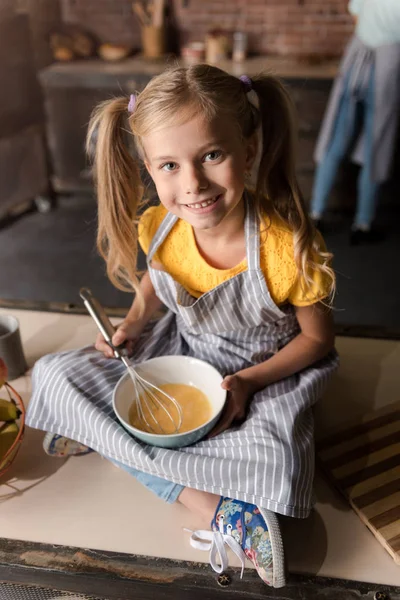 Fröhliches kleines Mädchen, das Eier mischt, während es auf dem Tisch sitzt — Stockfoto