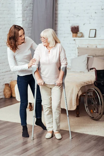 Mujer positiva ayudando a su abuela a caminar con muletas —  Fotos de Stock