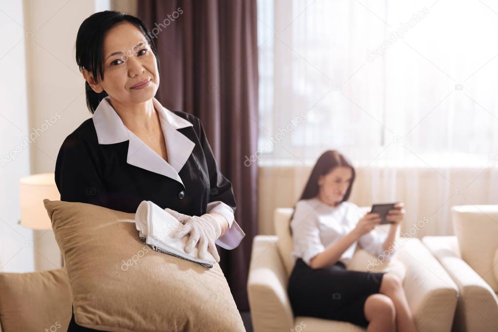 Positive good looking hotel maid cleaning the cushion