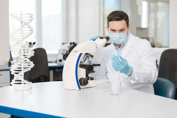 Человек ученый в маске сидит рядом с микроскопом — стоковое фото