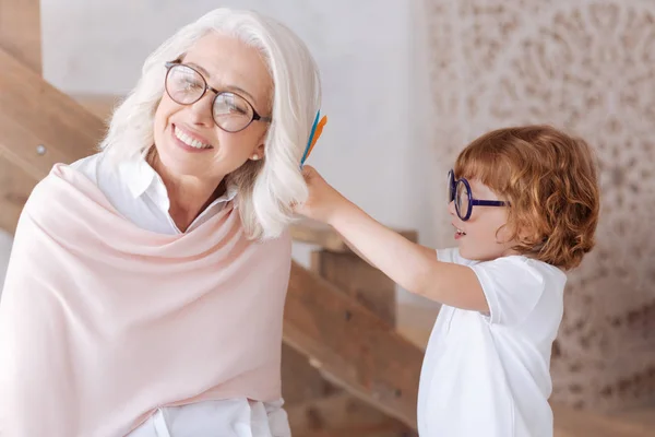 Cute pleasant boy putting feathers into his grandmothers hair — Stock Photo, Image