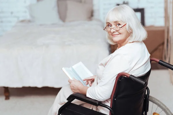 Mulher aposentada positiva ler livro em cadeira de rodas em casa — Fotografia de Stock