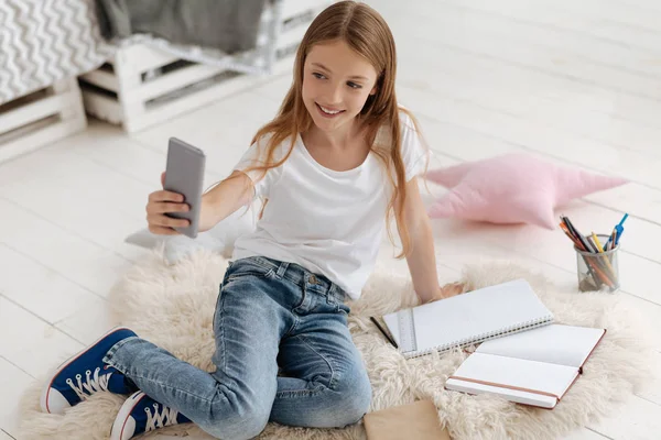 Menina radiante tomando auto retratos ao fazer lição de casa — Fotografia de Stock