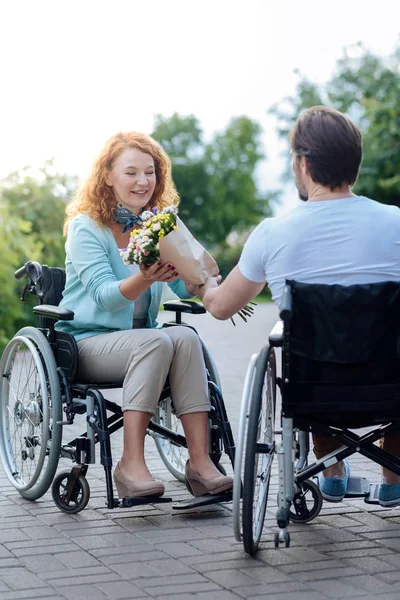 Mujer en silla de ruedas positiva recibiendo flores de su marido discapacitado cuidado — Foto de Stock