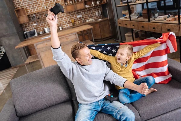 Rapaz alegre embrulhado em bandeira dos EUA aplaudindo com o pai — Fotografia de Stock