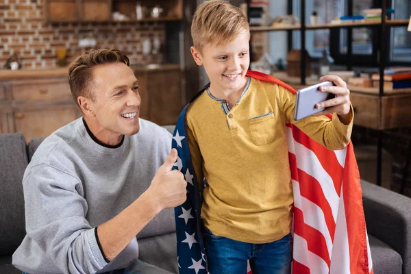 Menino adorável envolto em bandeira dos EUA tirando selfie com o pai — Fotografia de Stock
