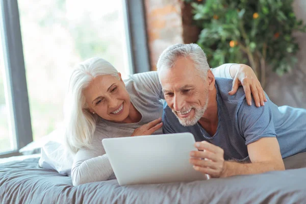 Casal idoso alegre descansando na cama com laptop — Fotografia de Stock