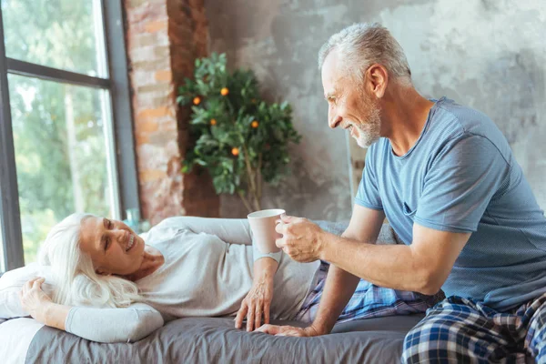 Homem idoso positivo trazendo um chá da manhã para sua esposa — Fotografia de Stock