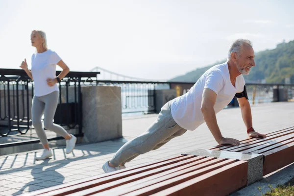 Senior man doing push-ups while his wife jogging — Stock Photo, Image