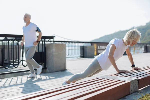 Elderly lady doing push-ups while her husband jogging — Stock Photo, Image