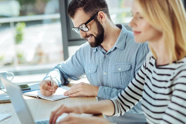 Uomo sorridente che scrive nel taccuino mentre collega lavora sul computer portatile — Foto Stock