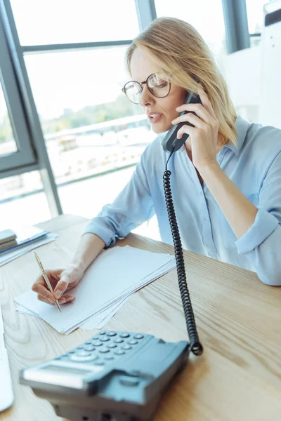Pretty businesswoman making notes while talking on phone — Stock Photo, Image