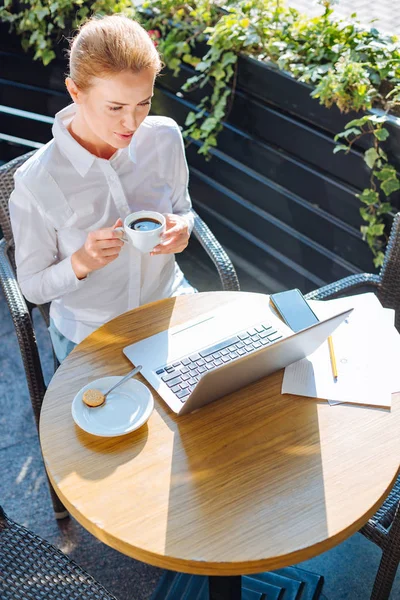 Красивая женщина пьет кофе во время чтения с ноутбука — стоковое фото