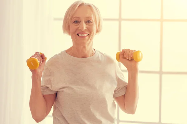 Positive senior woman exercising with dumbbells at home