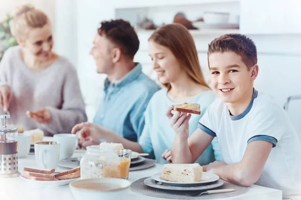 Щасливий молодший посміхається в камеру, снідаючи з сім'єю — стокове фото