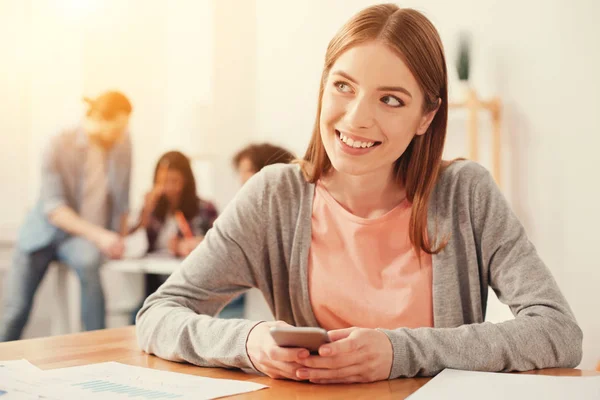 Usmívající se student sedí u stolu s chytrým telefonem v ruce — Stock fotografie