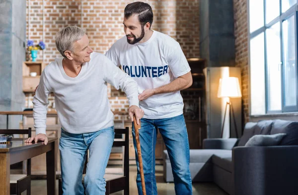 Trevlig typ volontär stödja en äldre man och hjälpa honom medan promenader — Stockfoto