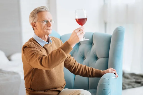 Homem velho concentrado usando óculos examinando vinho tinto — Fotografia de Stock