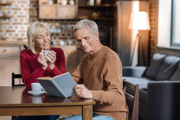 Веселый пенсионер читает книгу, пока его жена пьет чай. — стоковое фото
