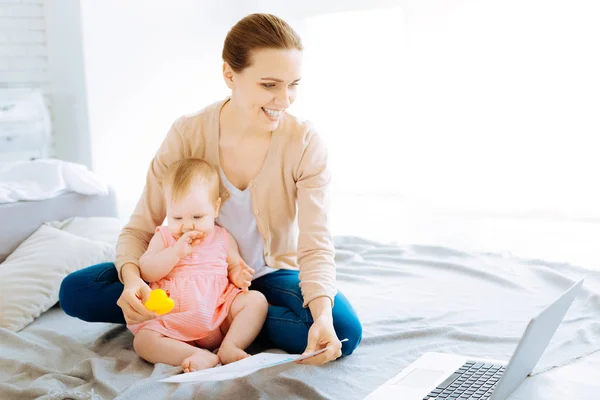 Cheerful mother sitting with a child on a bed while working — Stock Photo, Image
