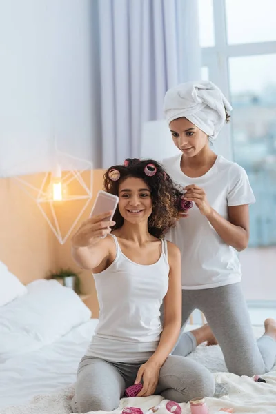 Sonriente chica tomando selfies mientras riza su cabello con su hermana —  Fotos de Stock