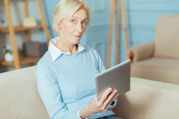 Neugierige alte Frau sitzt auf einem Sofa und hält ein modernes Tablet — Stockfoto