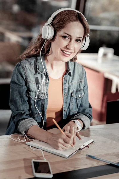 Mujer joven positiva escuchando música mientras toma notas —  Fotos de Stock