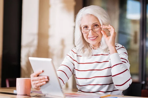 Mujer de edad avanzada inteligente mirando feliz mientras se sienta con un dispositivo moderno — Foto de Stock