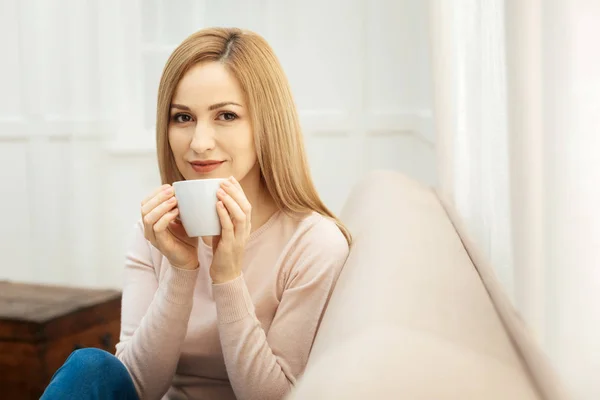 Mujer joven y bonita sentada en el sofá sosteniendo una taza — Foto de Stock