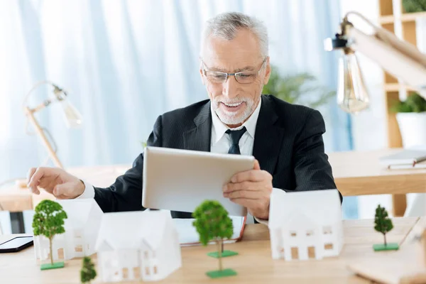 Positiver Arbeiter sitzt am Tisch und lächelt und hält ein Gerät in der Hand — Stockfoto