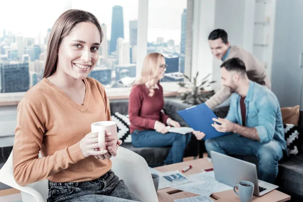 Piacevole donna sorridente che tiene una tazza di tè e seduta . — Foto Stock