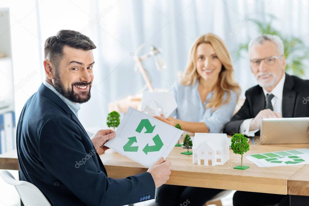 Happy man holding a recycling symbol while sitting with his colleagues