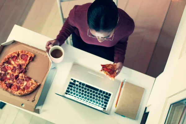 Ovanifrån av ung kvinna äter pizza medan du arbetar — Stockfoto