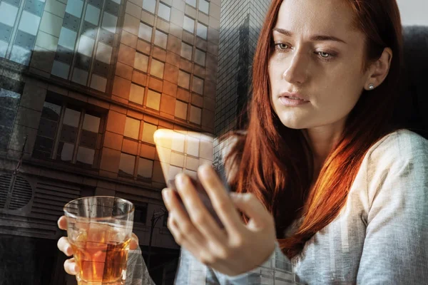 Serious woman feeling bad while drinking alcohol and looking at her smartphone — Stock Photo, Image
