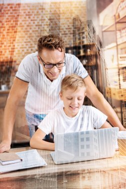 Babasının yanında ayakta iken bir dizüstü bilgisayar ile oturup boy gülümseyerek