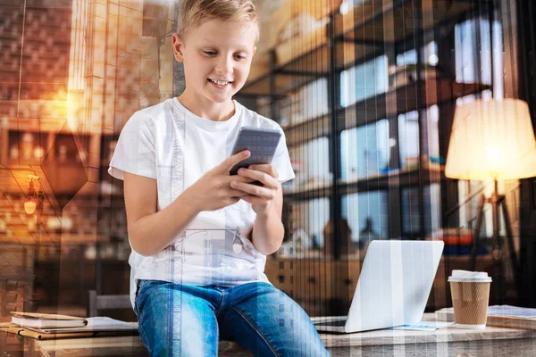 Счастливый мальчик сидит на столе и смотрит на свой смартфон — стоковое фото