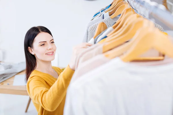 Mujer joven positiva sonriendo mientras mira la ropa nueva —  Fotos de Stock