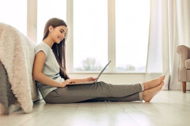 Pleasant teenage girl sitting on floor and using laptop clipart