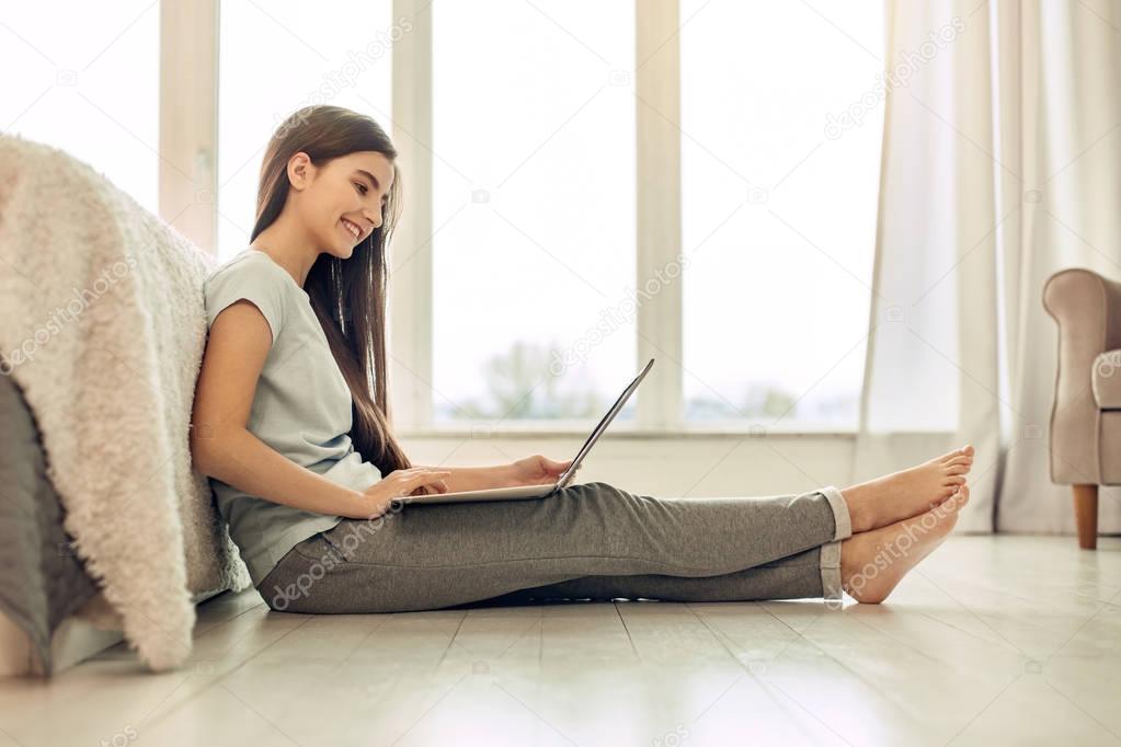 Pleasant teenage girl sitting on floor and using laptop